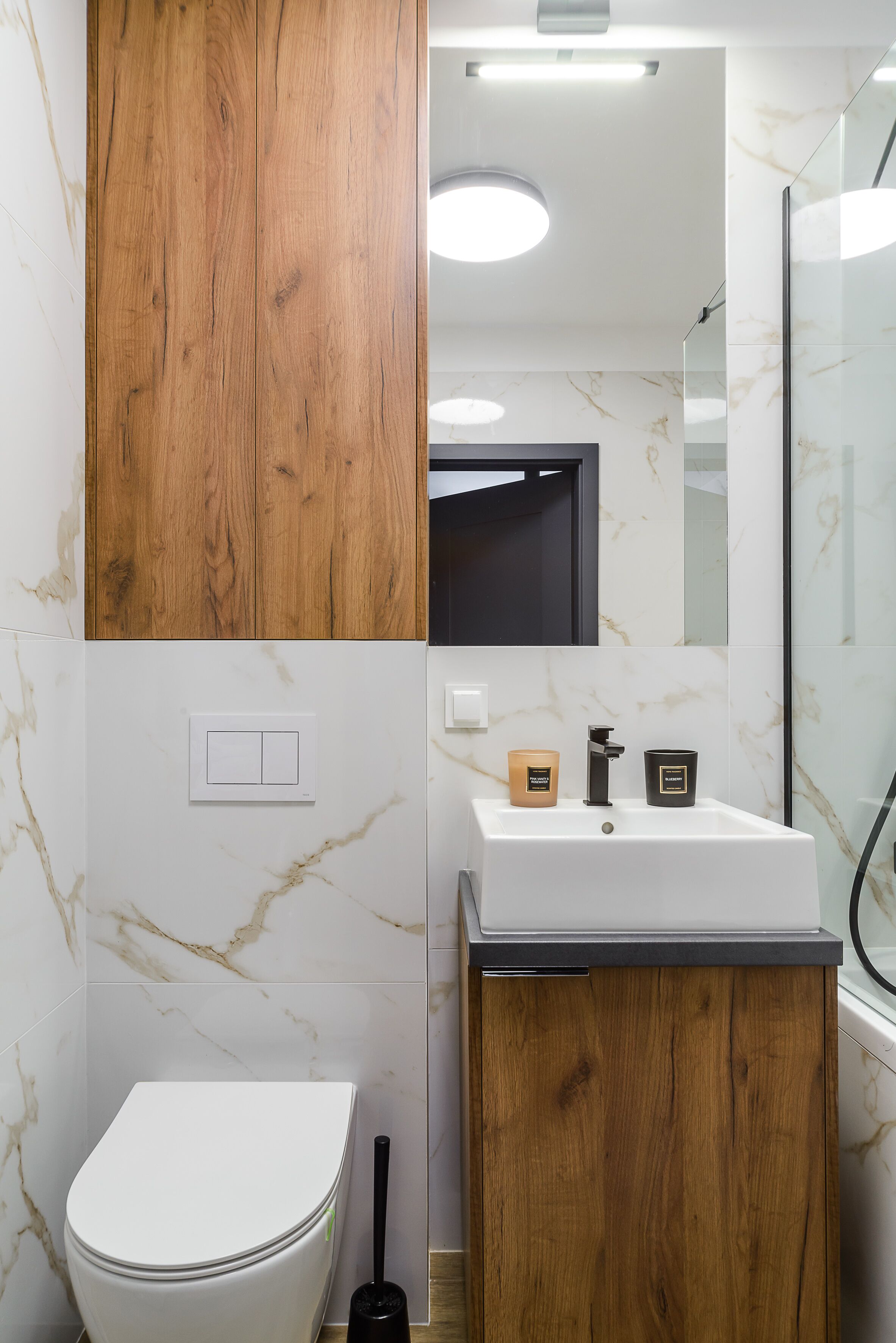 A bathroom with a white toilet, wooden vanity, marble tiles, a white sink, and a mirror.