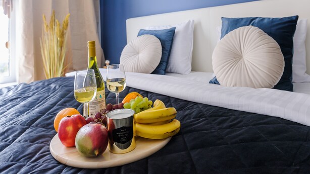 A bed with a dark blue bedspread, white pillows, and a wooden tray with fruits and a bottle of wine. - Lumina deluxe 3-room apartment (Lodz)