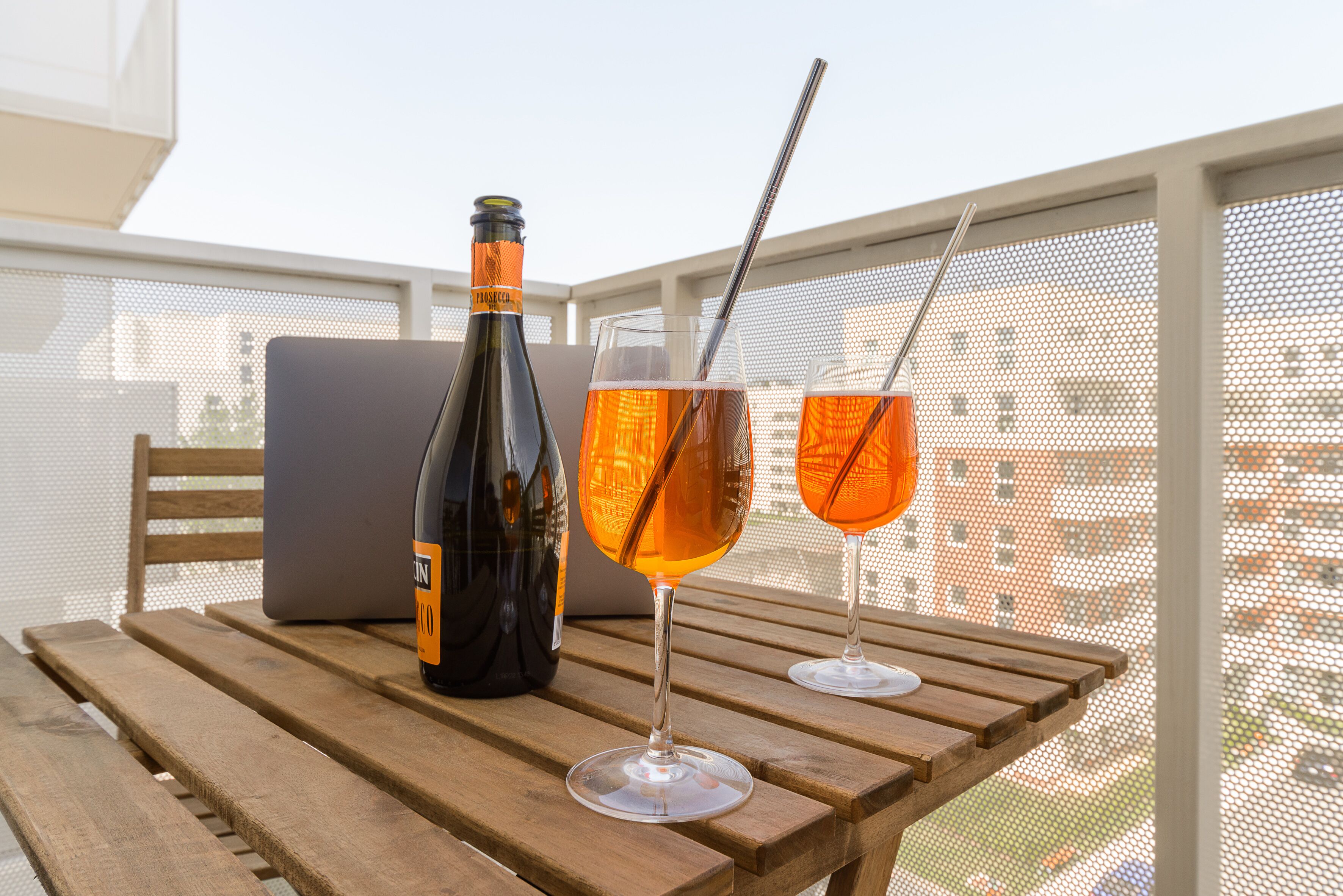 Una mesa de madera con una botella y dos vasos de líquido ámbar, una computadora portátil y una pantalla mosquitera al fondo.