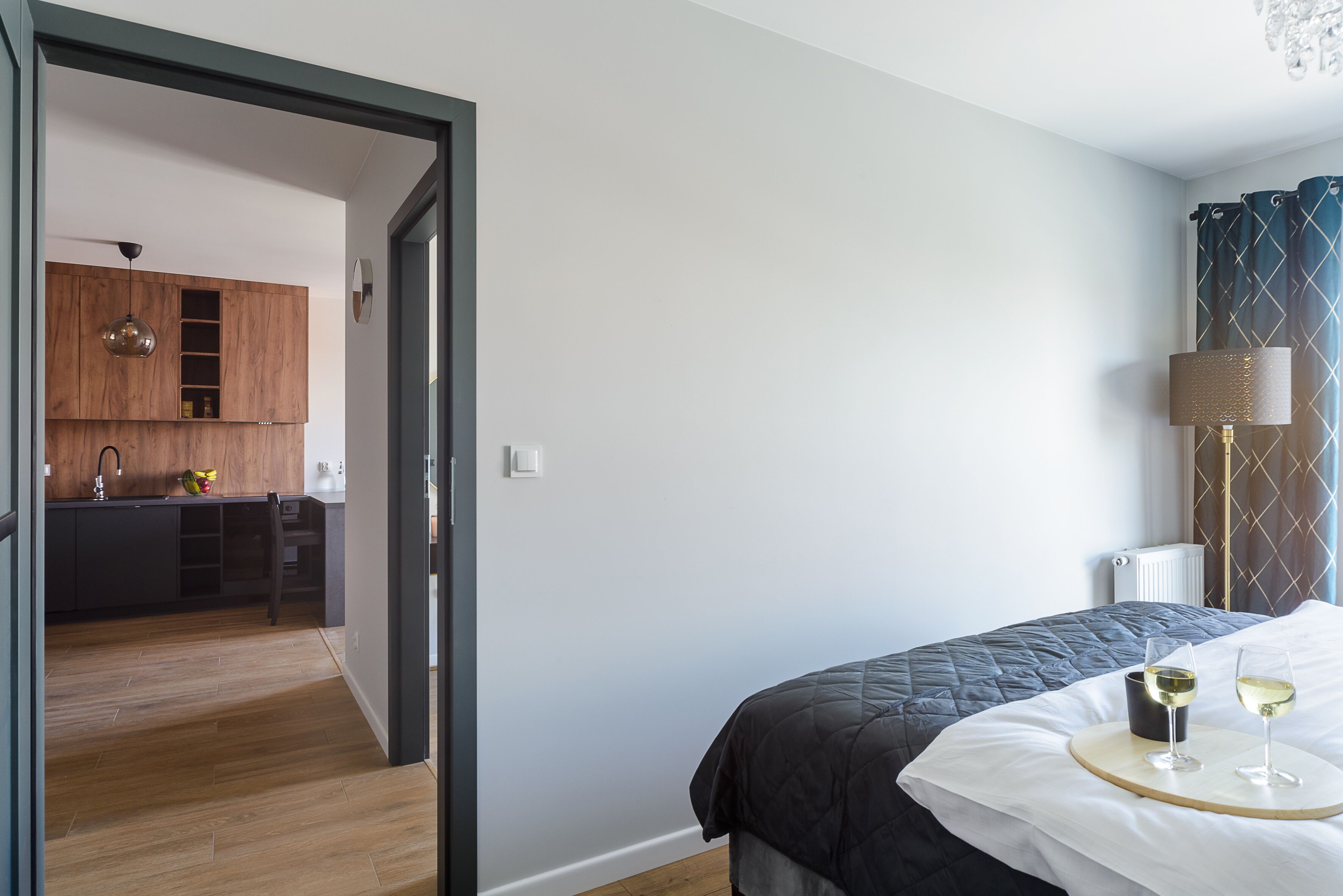 Ein Schlafzimmer mit einem Bett, einem Nachttisch mit Tablett und zwei Weingläsern sowie Blick in eine moderne Küche mit Holzschränken und einer Pendelleuchte.