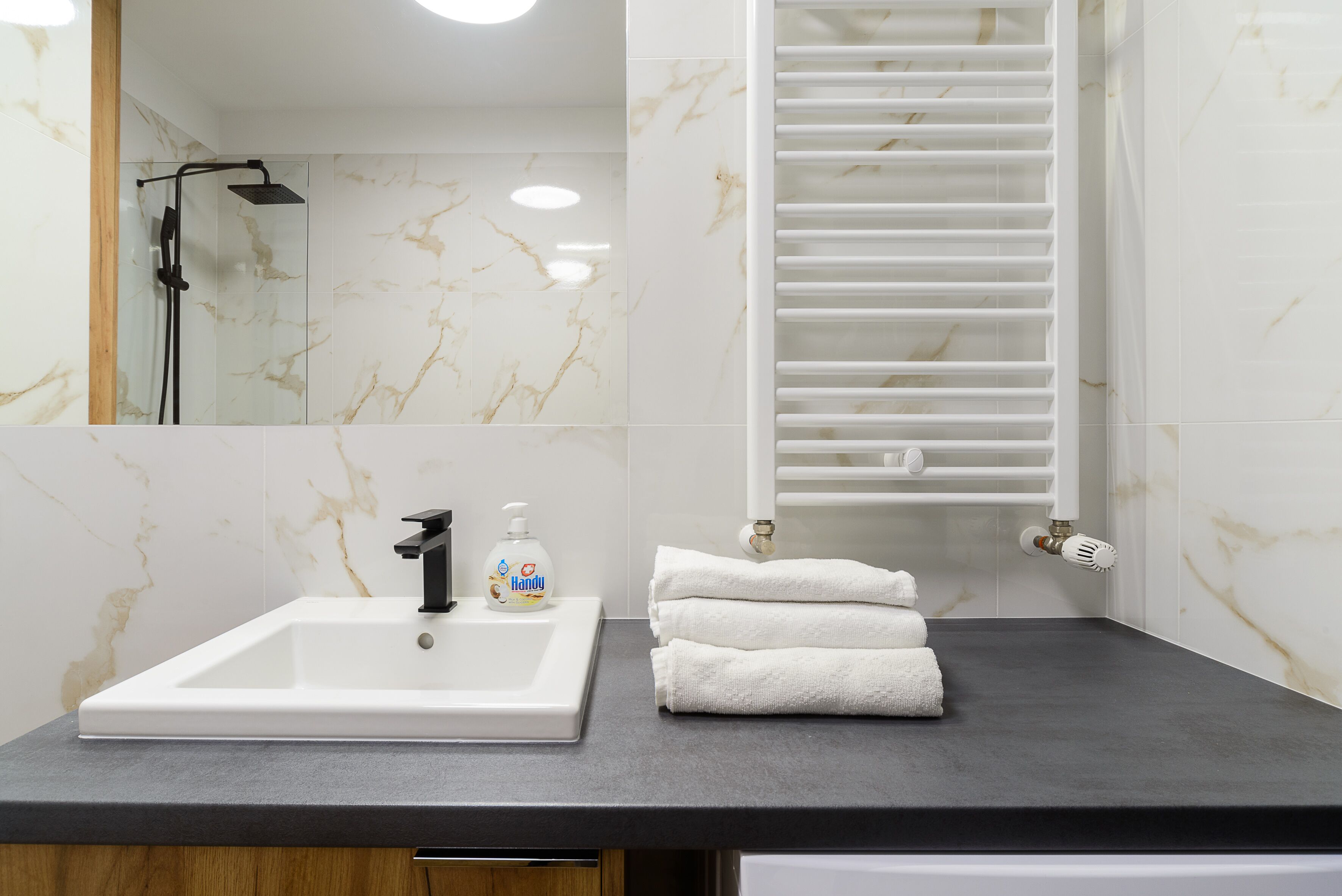 Une salle de bain avec un lavabo blanc, une douchette à main, un porte-serviettes avec des serviettes et un flacon de savon liquide.