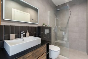 Uma casa de banho moderna com uma grande pia branca, um revestimento de azulejos escuros, uma cabine de chuveiro de vidro e uma sanita.