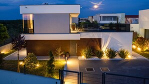 A modern two-story house with a flat roof, large windows, and a well-lit outdoor area with plants and a paved driveway. - Villa Marli (Nin)