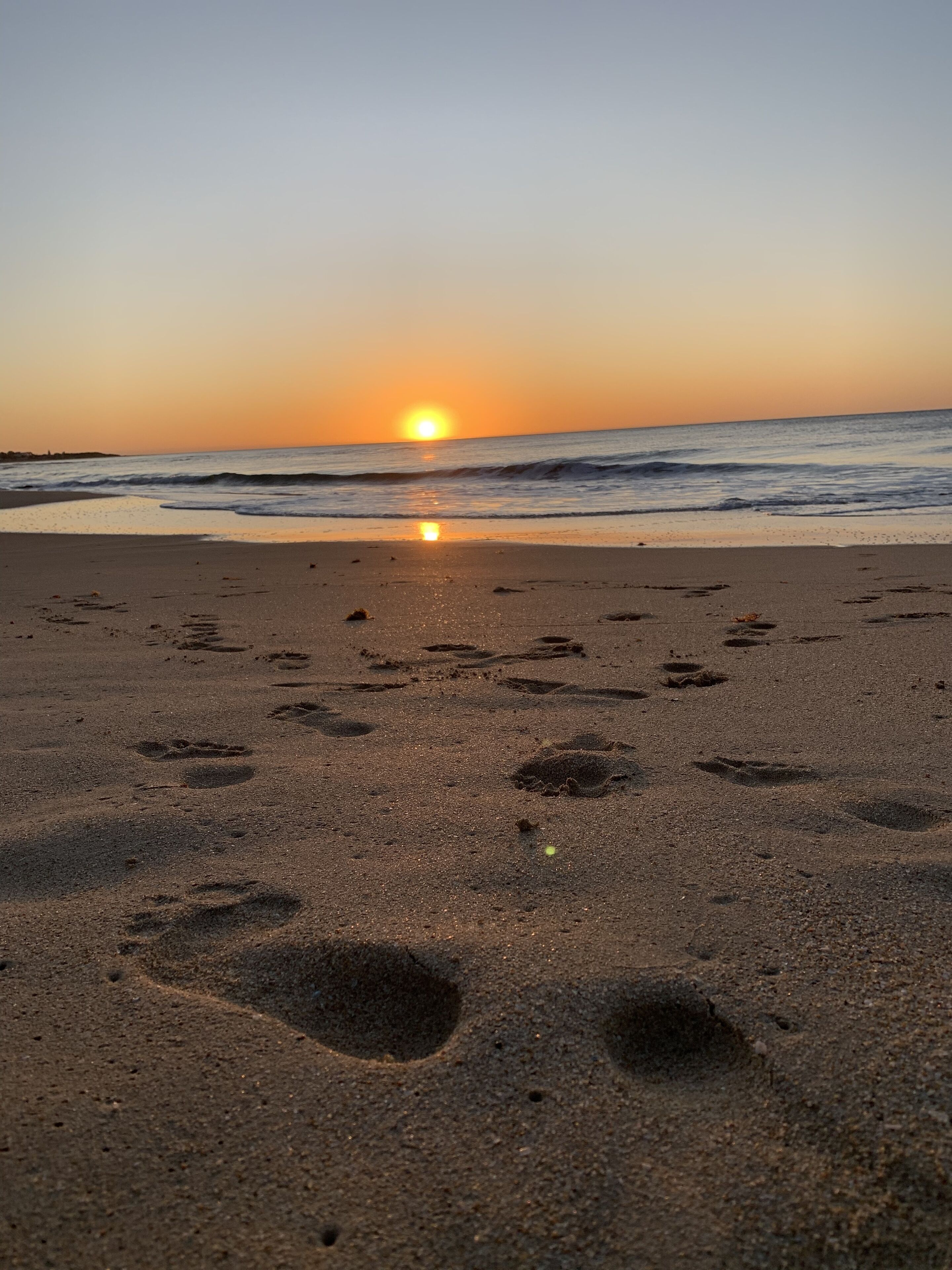 Ein Strand mit Fußspuren, die zum Wasser führen, ein Sonnenuntergang im Hintergrund und ein ruhiges Meer.