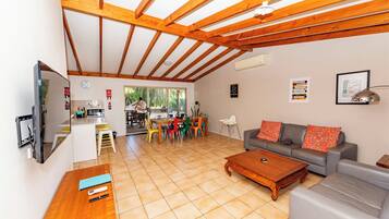 A spacious living area with a wooden ceiling, a grey sofa, a wooden coffee table, and a dining area with colorful chairs.