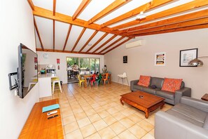 Een ruime woonkamer met een houten plafond, een grijze bank, een houten salontafel en een eethoek met kleurrijke stoelen.