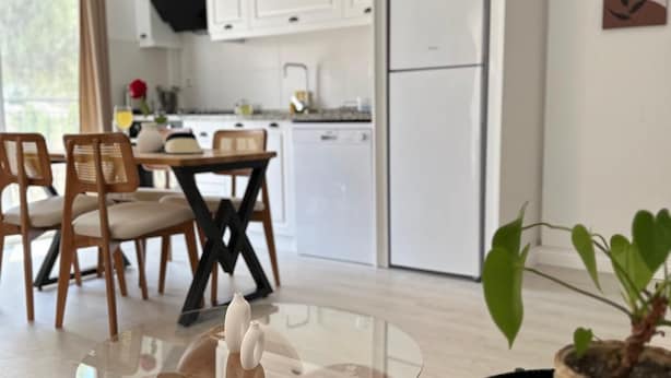 A modern kitchen with white cabinets, a dining area with wooden chairs, and a glass coffee table. - Likya Apart Kaş (Kas)