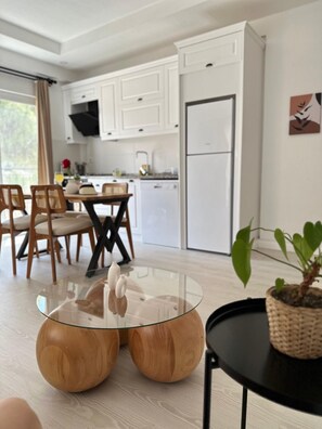 A modern kitchen with white cabinets, a dining area with wooden chairs, and a glass coffee table. - Likya Apart Kaş (Kas)