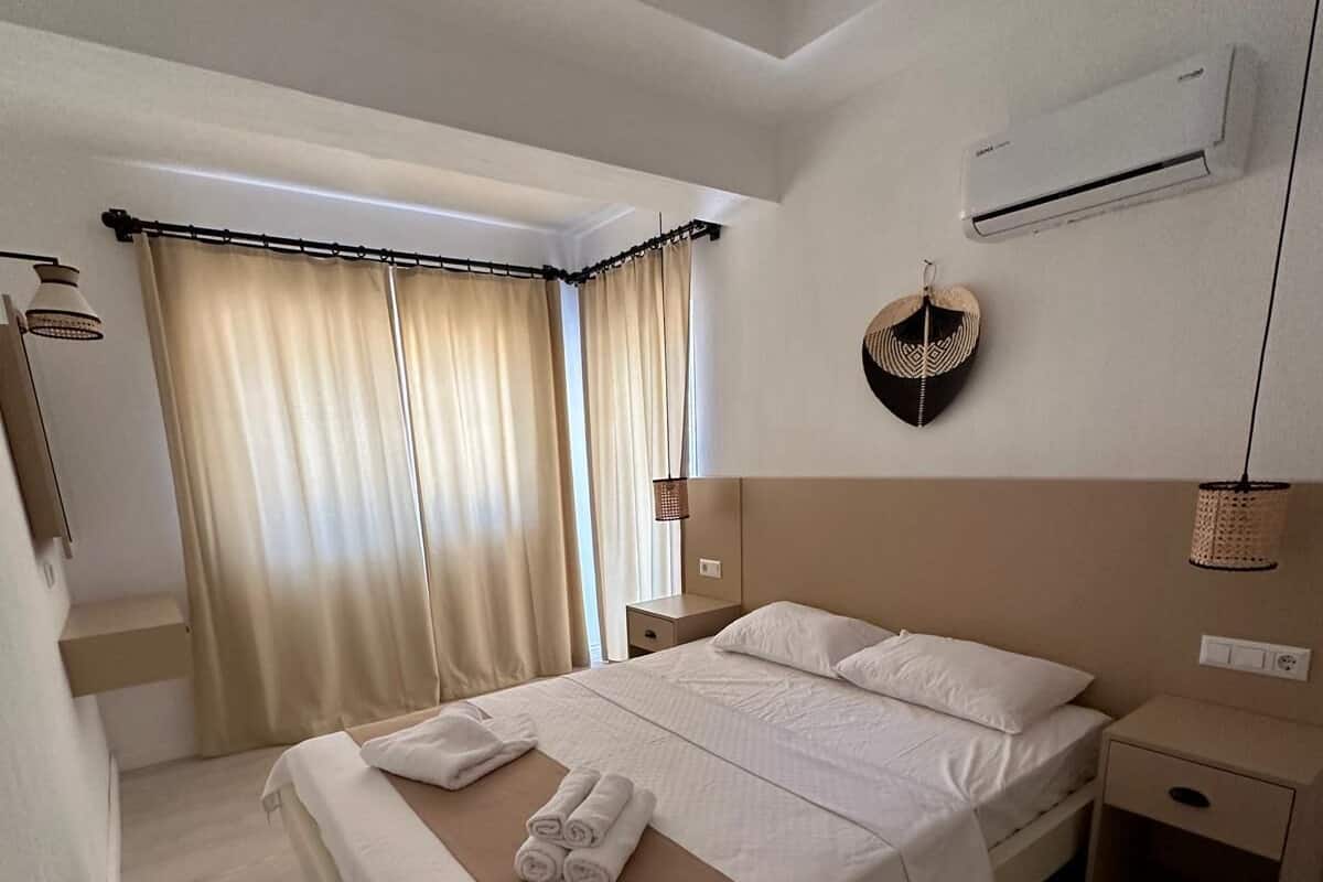A hotel room with a large bed, a bedside table, a wall-mounted air conditioner, and a window with curtains.
