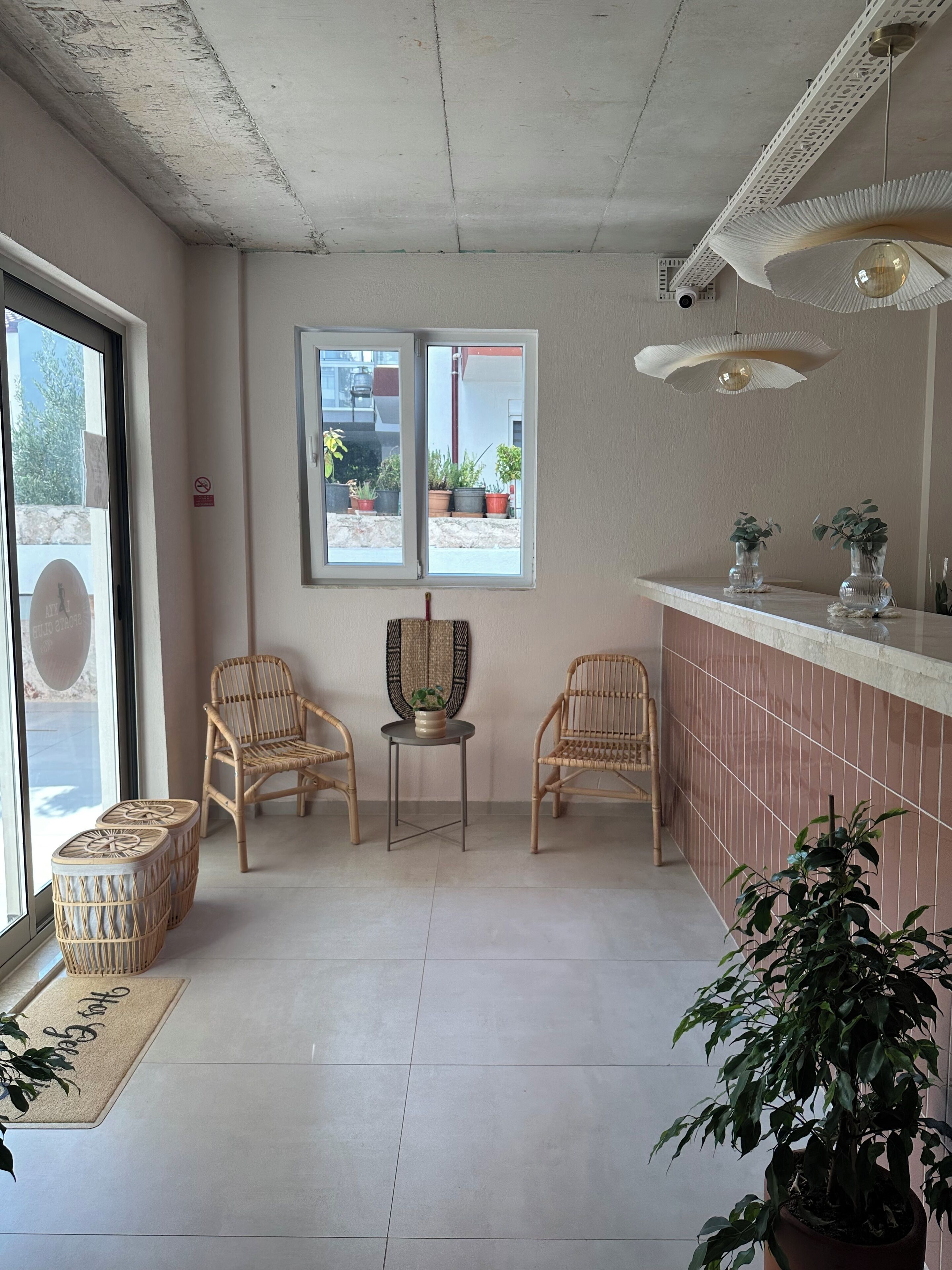 A room with two wicker chairs, a small table, a potted plant, a tiled floor, and a window with a view of the outside.