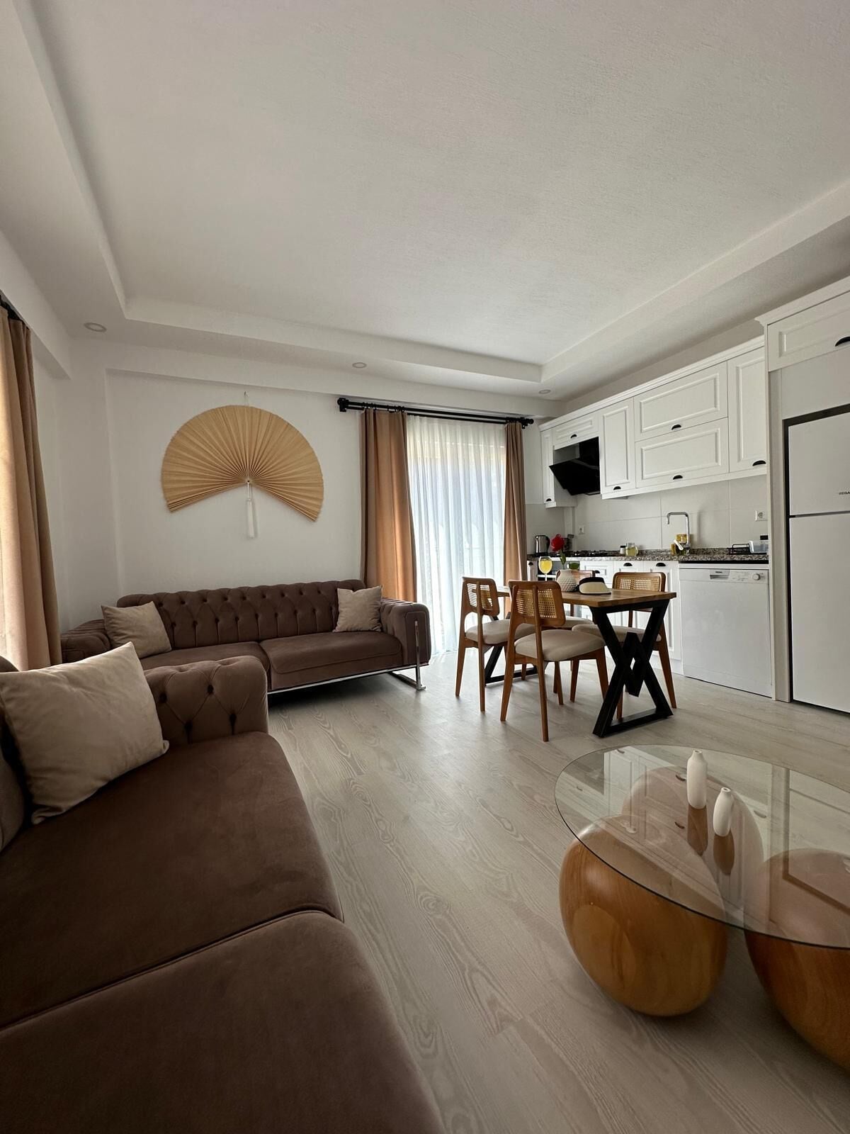 A modern living room with a brown sofa, a dining table with chairs, and a kitchen area with white cabinets and appliances.