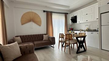 A modern living room with a brown sofa, a dining table with chairs, and a kitchen area with white cabinets and appliances.
