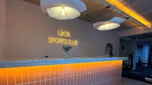 A reception area with a counter, a neon sign reading 'LIKYA SPORTS CLUB,' and a wicker chair. - Likya Apart Kaş (Kas)