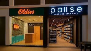Il y a un magasin appelé « Oldies », avec un comptoir en bois et des étagères remplies de produits. À côté, on trouve « Pallise Your One Stop Shop », qui propose une grande variété d’articles sur ses étagères.