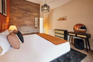 Ein modernes Schlafzimmer mit einer Wand in Holzoptik, einem Bett mit weißer Matratze und brauner Tagesdecke, einem Nachttisch mit Spiegel und einer Pendelleuchte.