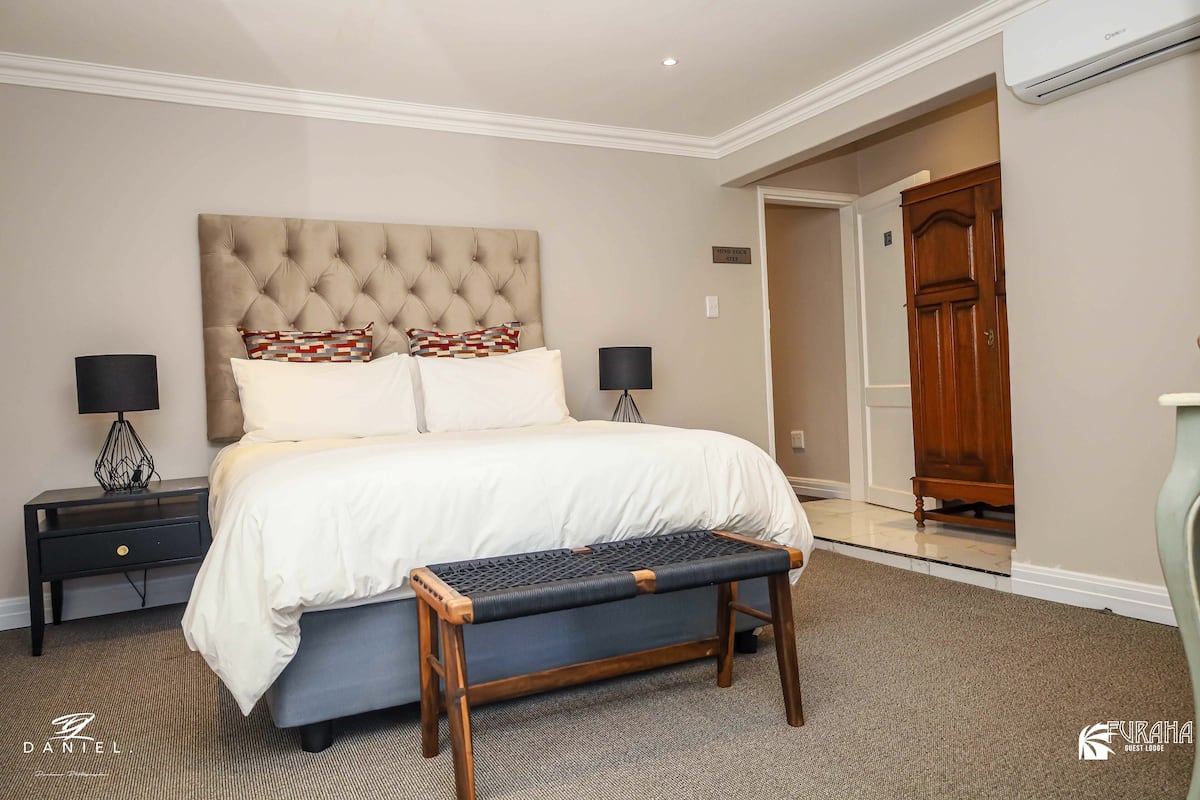 A bedroom with a tufted headboard, a large bed with white bedding, a bench with a woven seat, two bedside tables with lamps, and a wooden wardrobe.