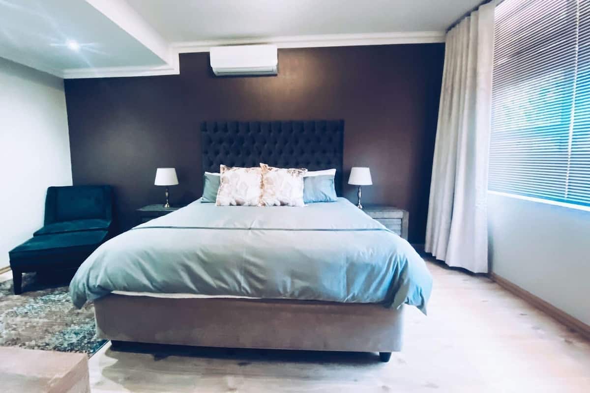 A modern bedroom with a large bed, two bedside tables, a chair, and a window with blinds.