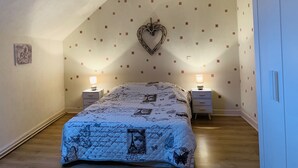A bedroom with a sloped ceiling, a heart-shaped wall decoration, a bed with a quilted cover, two bedside tables with lamps, and a door on the right.