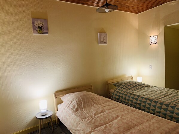 A room with two single beds, a small table with a lamp, and a wall-mounted light fixture.