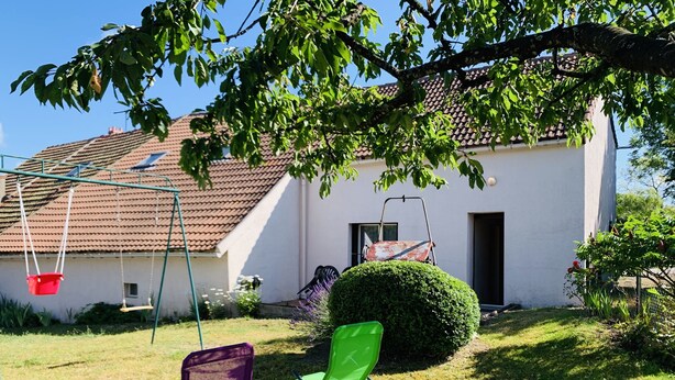 A white house with a red-tiled roof, a swing set, and two chairs in the yard.