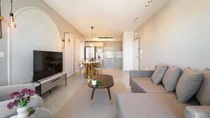 A modern living room with a flat-screen TV, a grey sofa, a coffee table, and a dining area with a chandelier.