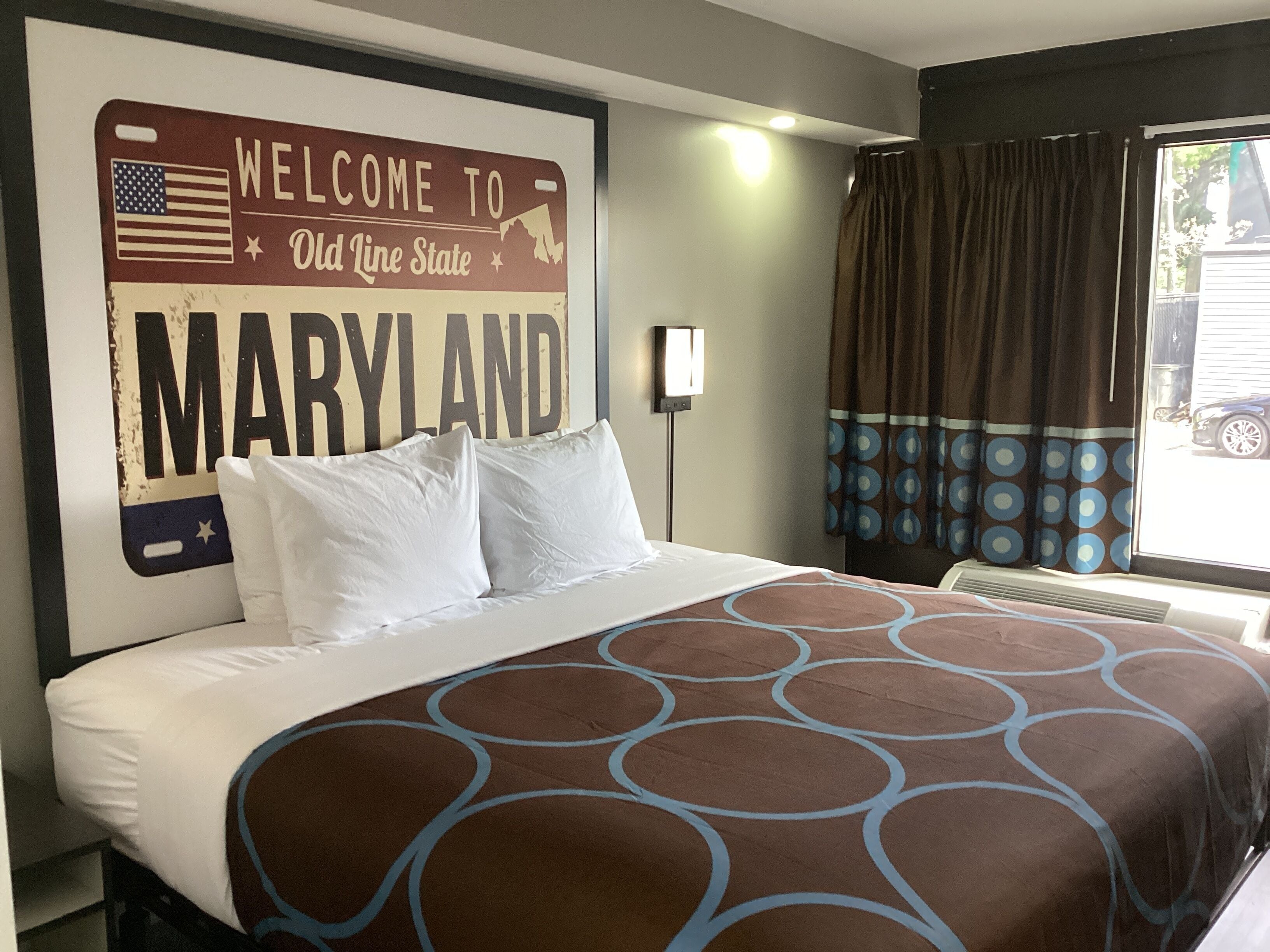 A hotel room with a large wall sign reading "Welcome to Maryland," a bed with white linens, and a window with curtains.