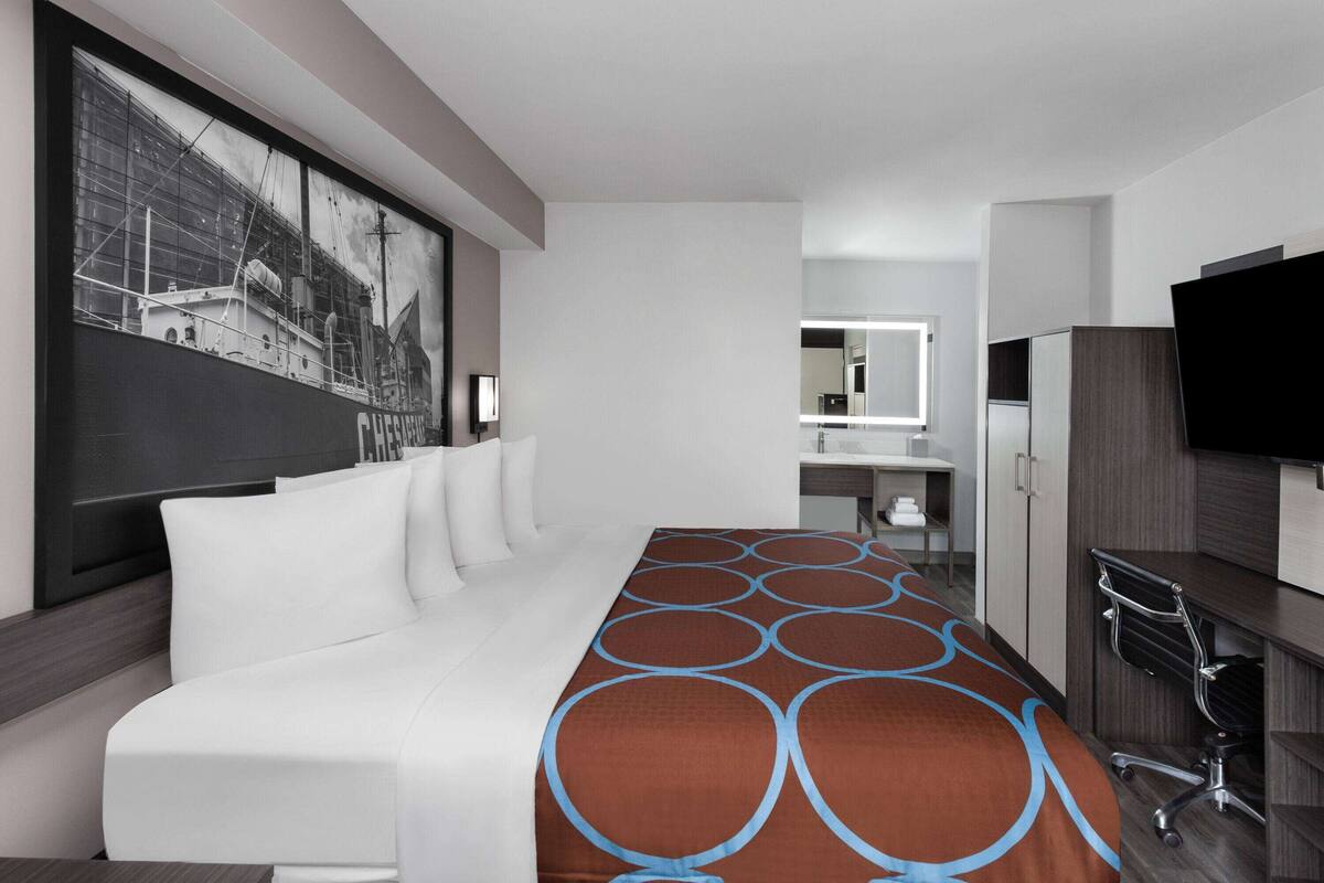 Moderni hotellihuone, jossa on suuri sänky, työpöytä tuolilla, televisio ja taustalla näkyvä kylpyhuone.