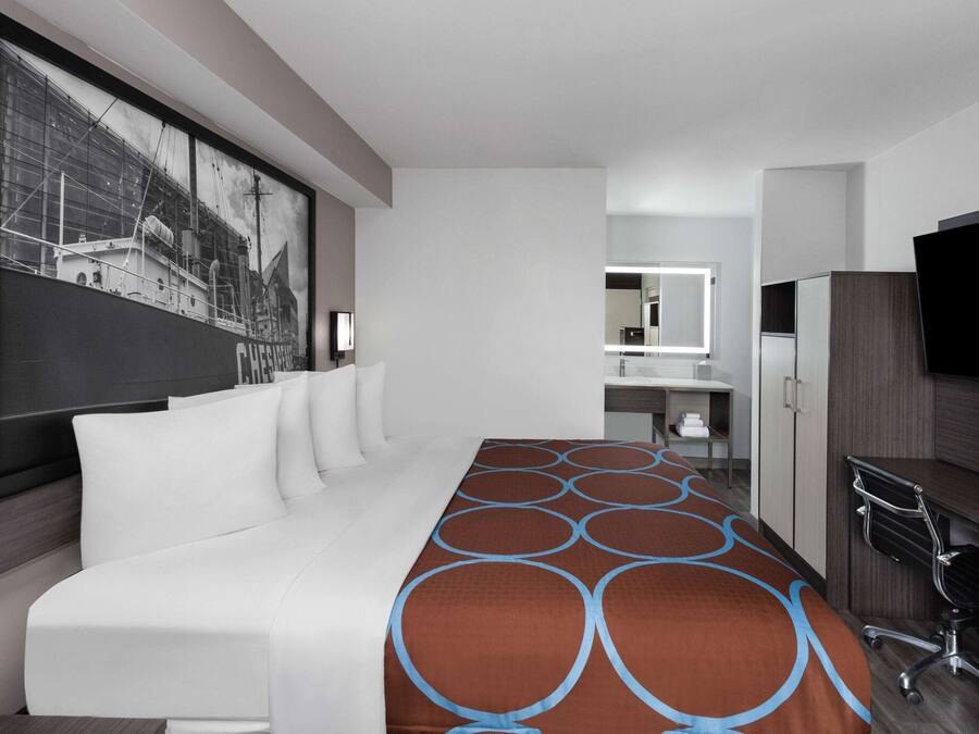 A modern hotel room with a large bed, a desk with a chair, a television, and a bathroom visible in the background.