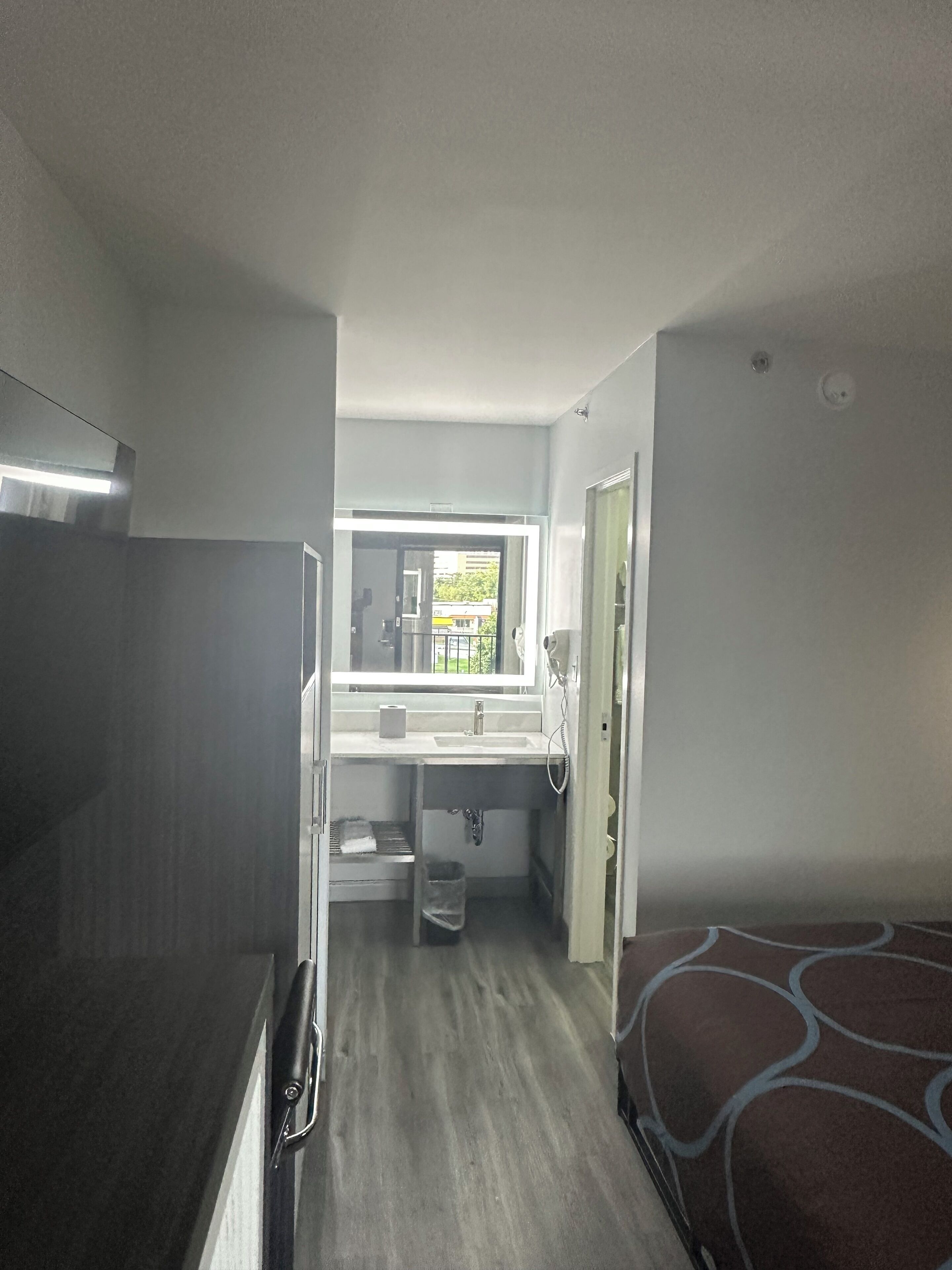 Habitación de hotel pequeña y funcional con kitchenette, baño reducido, ventana con vistas y una cama.