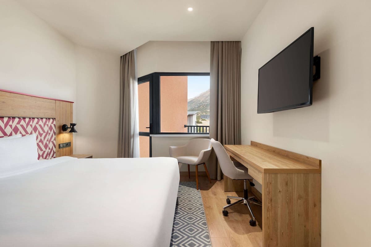 Une chambre d’hôtel comprenant un lit, un bureau, une chaise, un téléviseur fixé au mur et une fenêtre avec des rideaux.