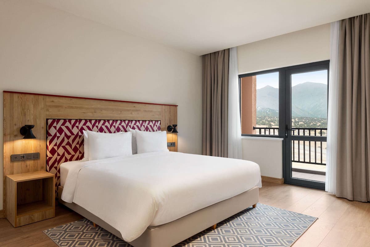Une chambre d’hôtel avec un grand lit, une tête de lit en bois, deux tables de chevet, un tapis à motifs et un balcon offrant une vue sur la montagne.