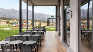 Un patio exterior con mesas y sillas, amplias ventanas que ofrecen vistas a parques infantiles y montañas.