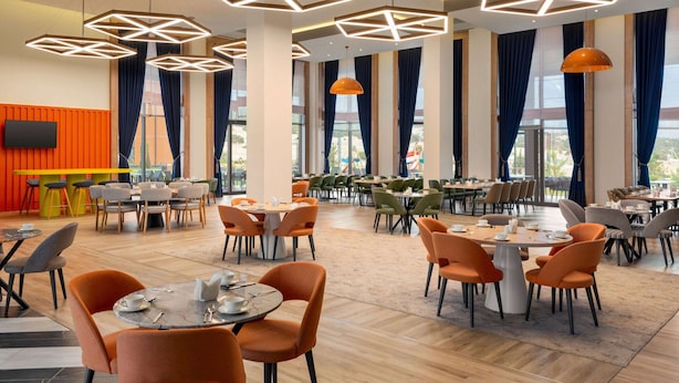 Un comedor amplio con varias mesas y sillas, luces de techo geométricas, ventanas grandes con cortinas y un televisor de pantalla plana montado en la pared.