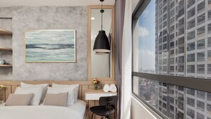 Una habitación de hotel moderna con una cama grande, un escritorio, una silla, un cuadro, un espejo y vistas a un paisaje urbano a través de un ventanal.