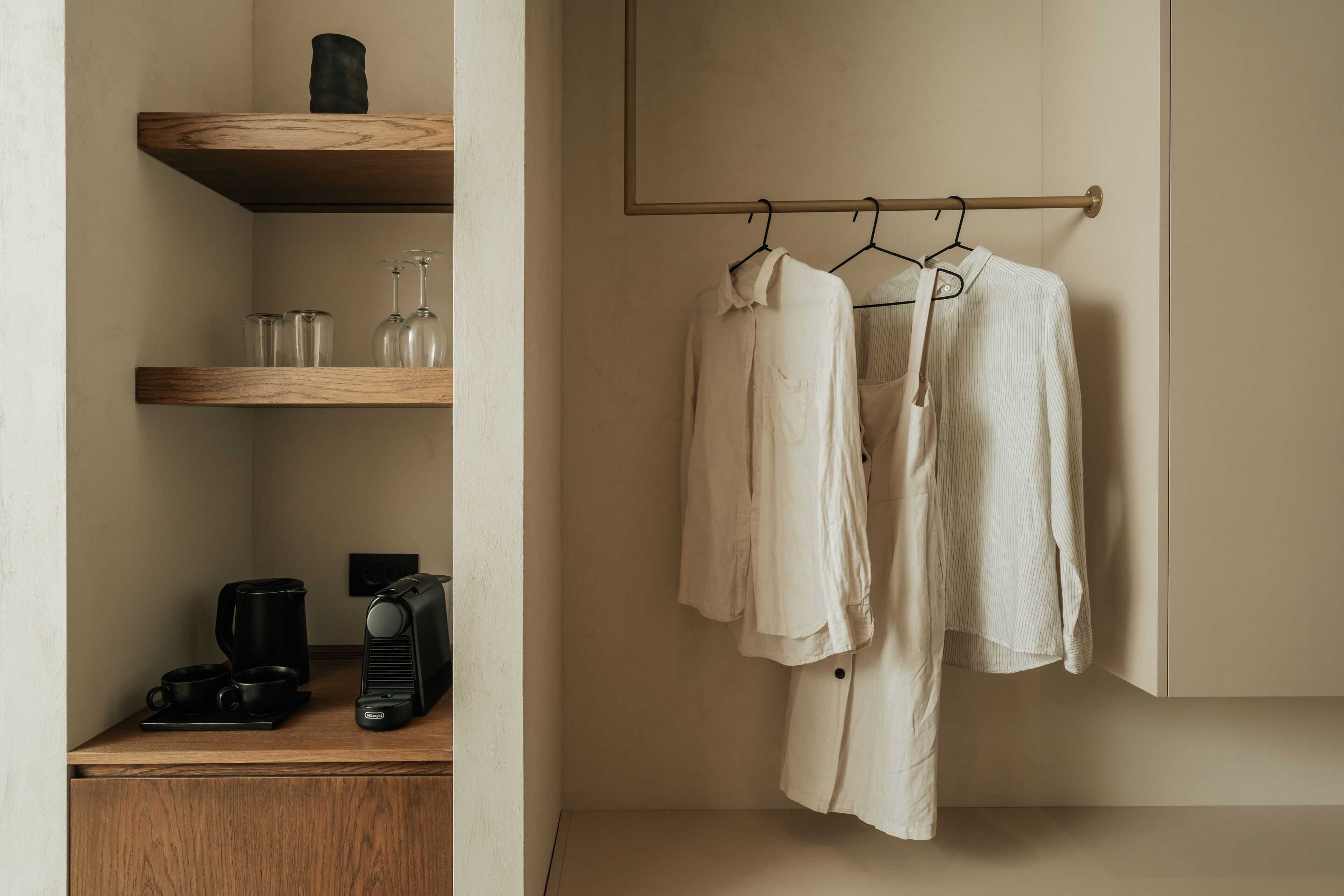 Habitación minimalista con una estantería de madera que sostiene vasos, una cafetera y un hervidor. Ropa colgada en un perchero adosado a la pared.