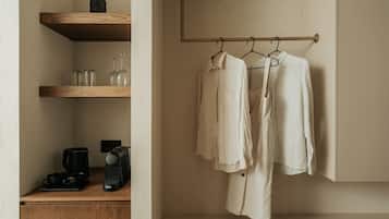 Habitación minimalista con una estantería de madera que sostiene vasos, una cafetera y un hervidor. Ropa colgada en un perchero adosado a la pared.