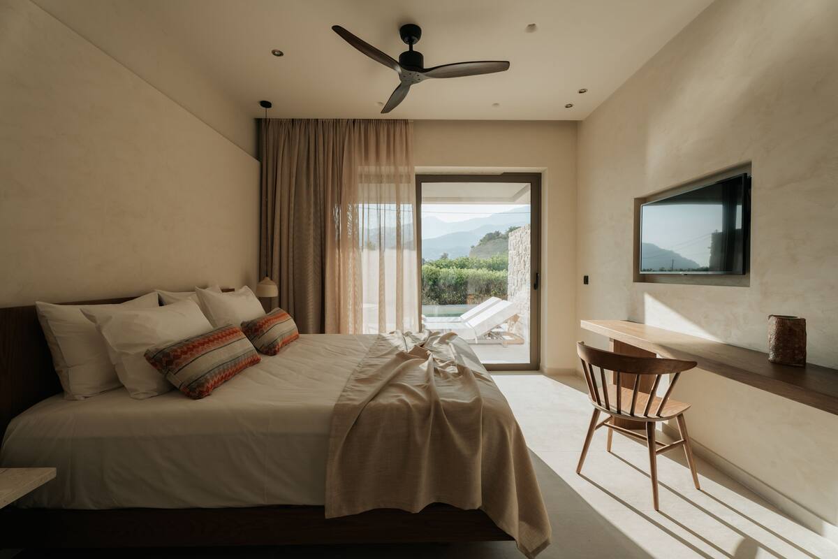 A modern bedroom with a bed, a ceiling fan, a TV, and a view of a pool and mountains.
