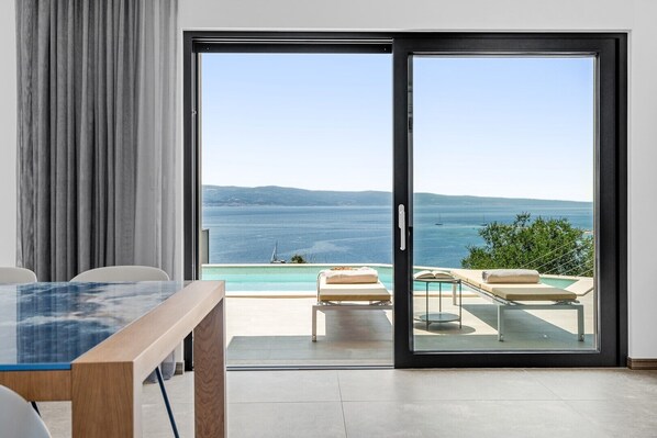 Une chambre moderne dotée d’une porte vitrée donnant sur un balcon avec une piscine et des chaises longues, offrant une vue panoramique sur l’océan.