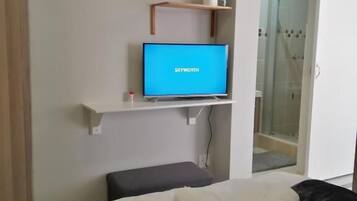 Quarto de hotel com uma cama, televisão em uma estante, área de chuveiro e um pequeno banco.