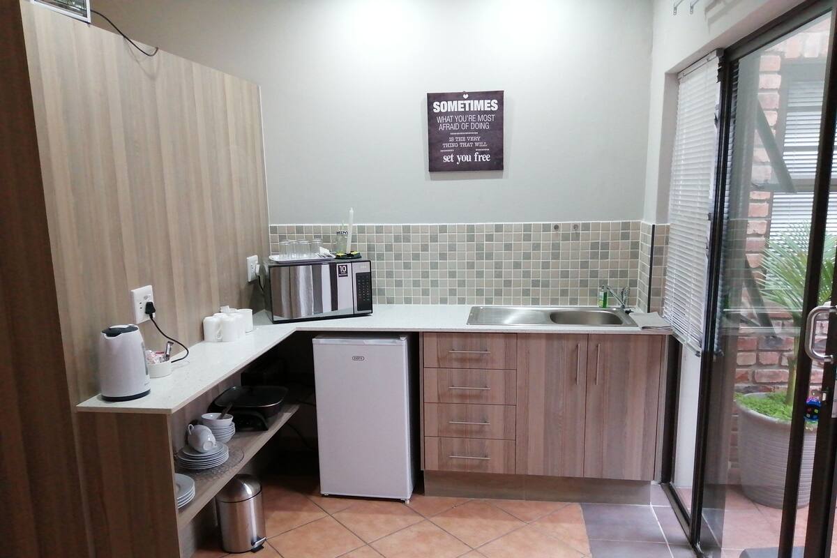 Una cucina compatta con frigorifero, microonde e lavandino. Sul muro c'è un cartello e una finestra con persiane.