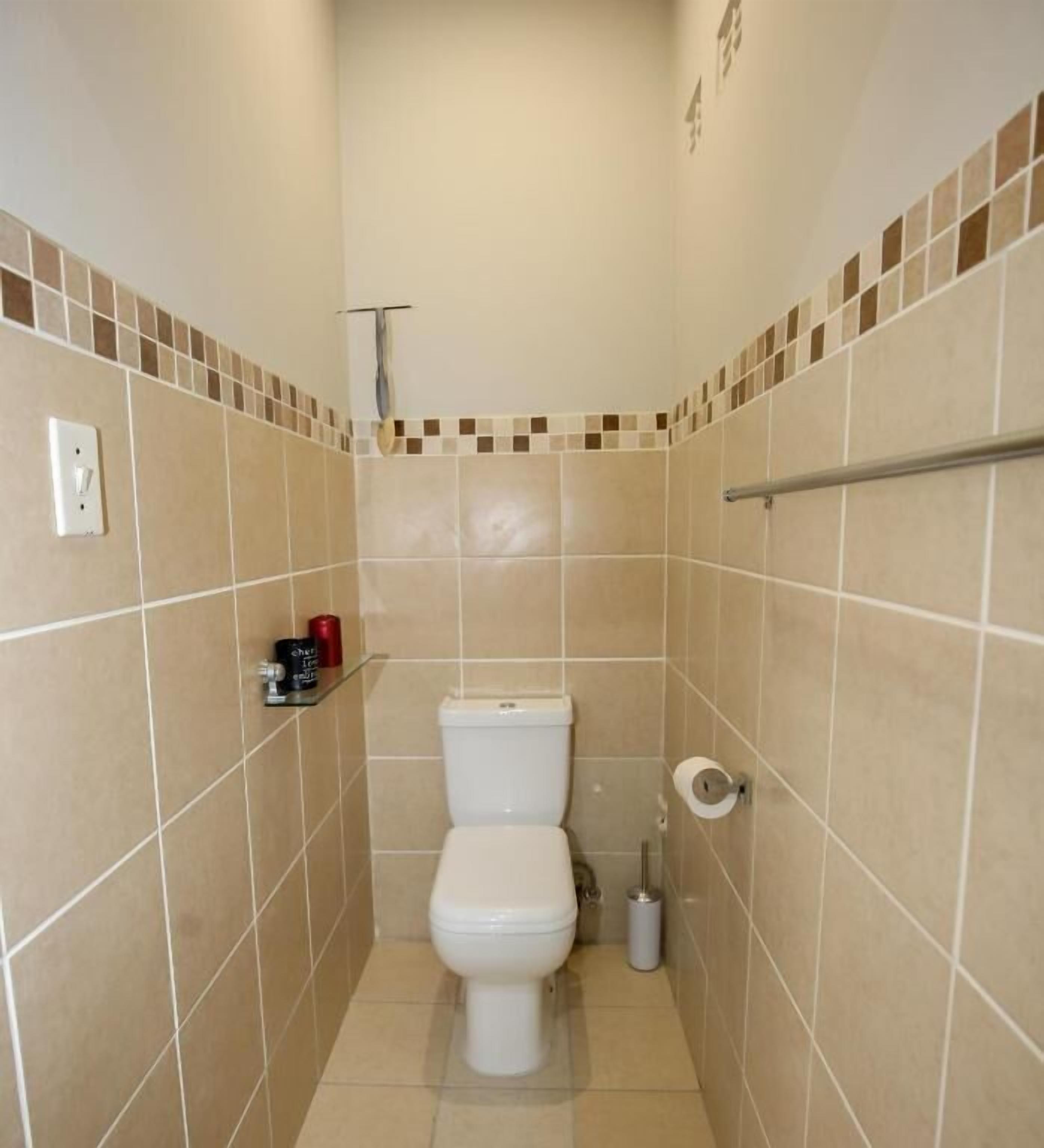 A small, narrow bathroom with beige tiles, a white toilet, a hand dryer, a toilet paper holder, and a small shelf with toiletries.