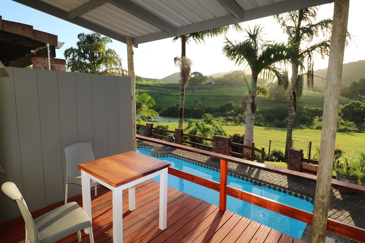 Un terrazzo in legno con piscina, sedie bianche e un tavolo con vista su un paesaggio lussureggiante con palme e colline.