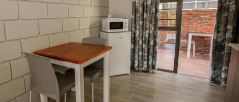 A small room with a wooden table and a white chair, a microwave on a small white cabinet, and a sliding glass door leading to an outdoor area with a brick wall and a table.