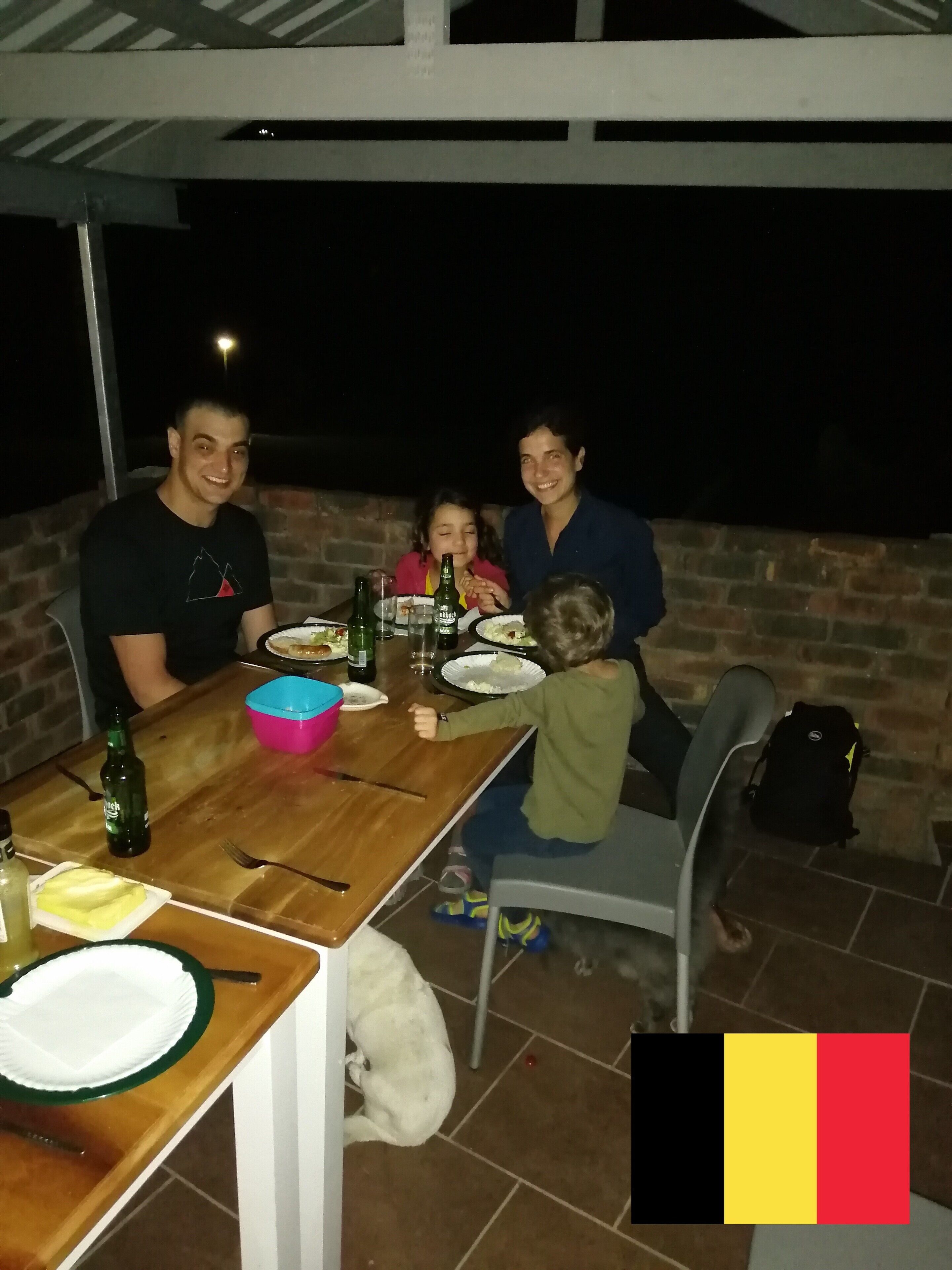 A family dining at an outdoor table with a brick wall backdrop, a dog on the floor, and a Belgian flag in the corner.