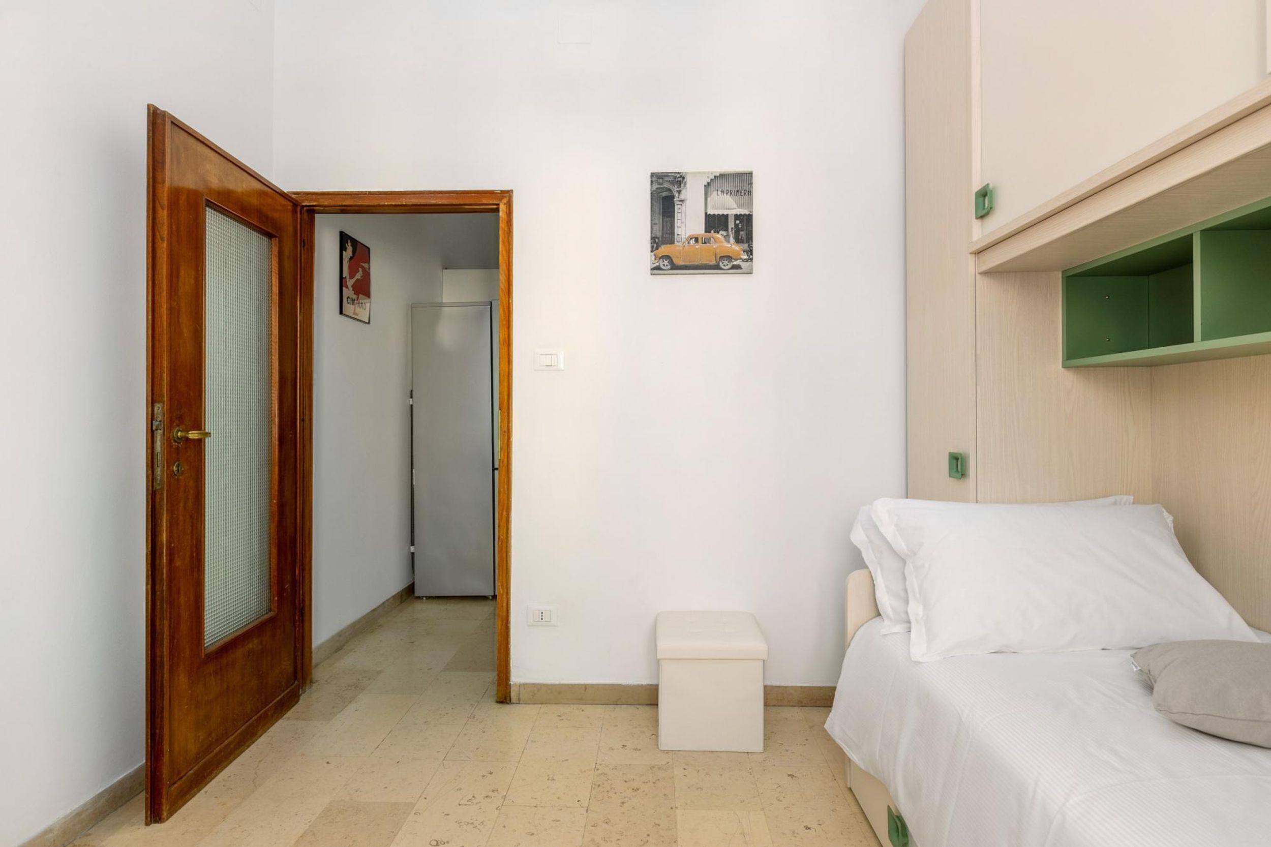 A small, clean room with a bed, a wooden door, a small white stool, and a green shelf.
