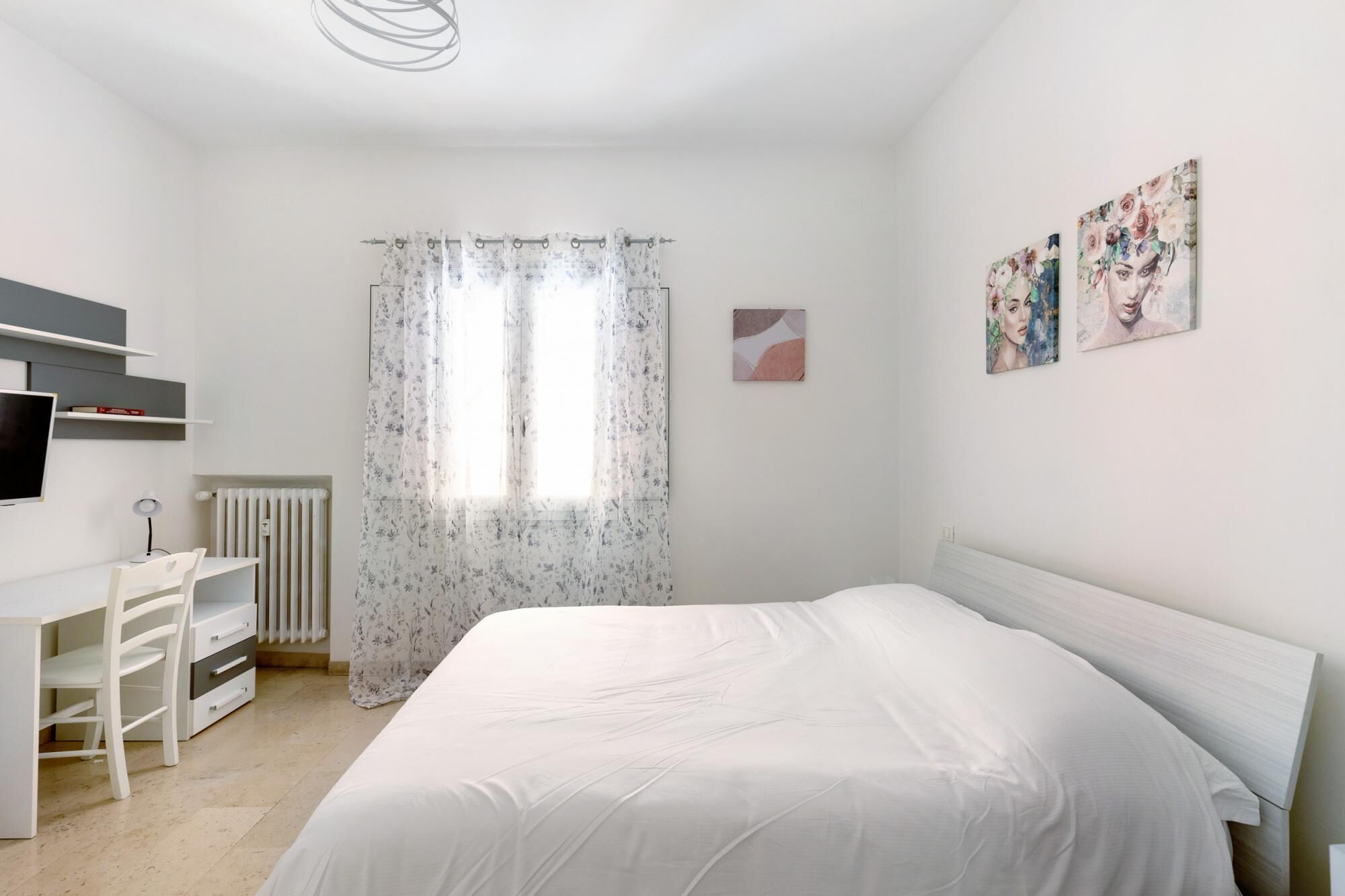 Une chambre à coucher comprenant un lit, un bureau, une chaise, un radiateur et une fenêtre avec des rideaux à motifs floraux.