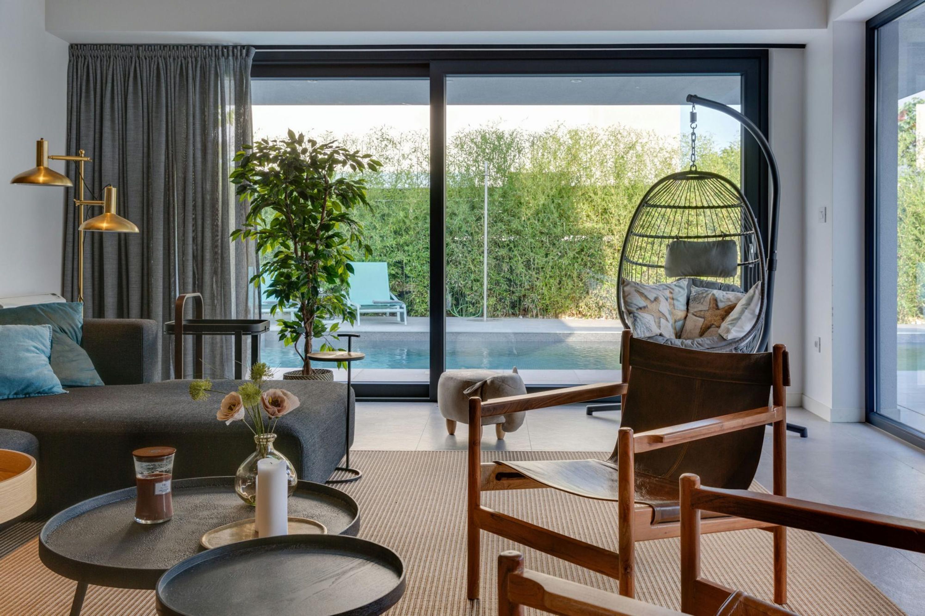 A modern living room with a sofa, a hanging chair, a small table with candles, and a view of a pool and greenery outside.