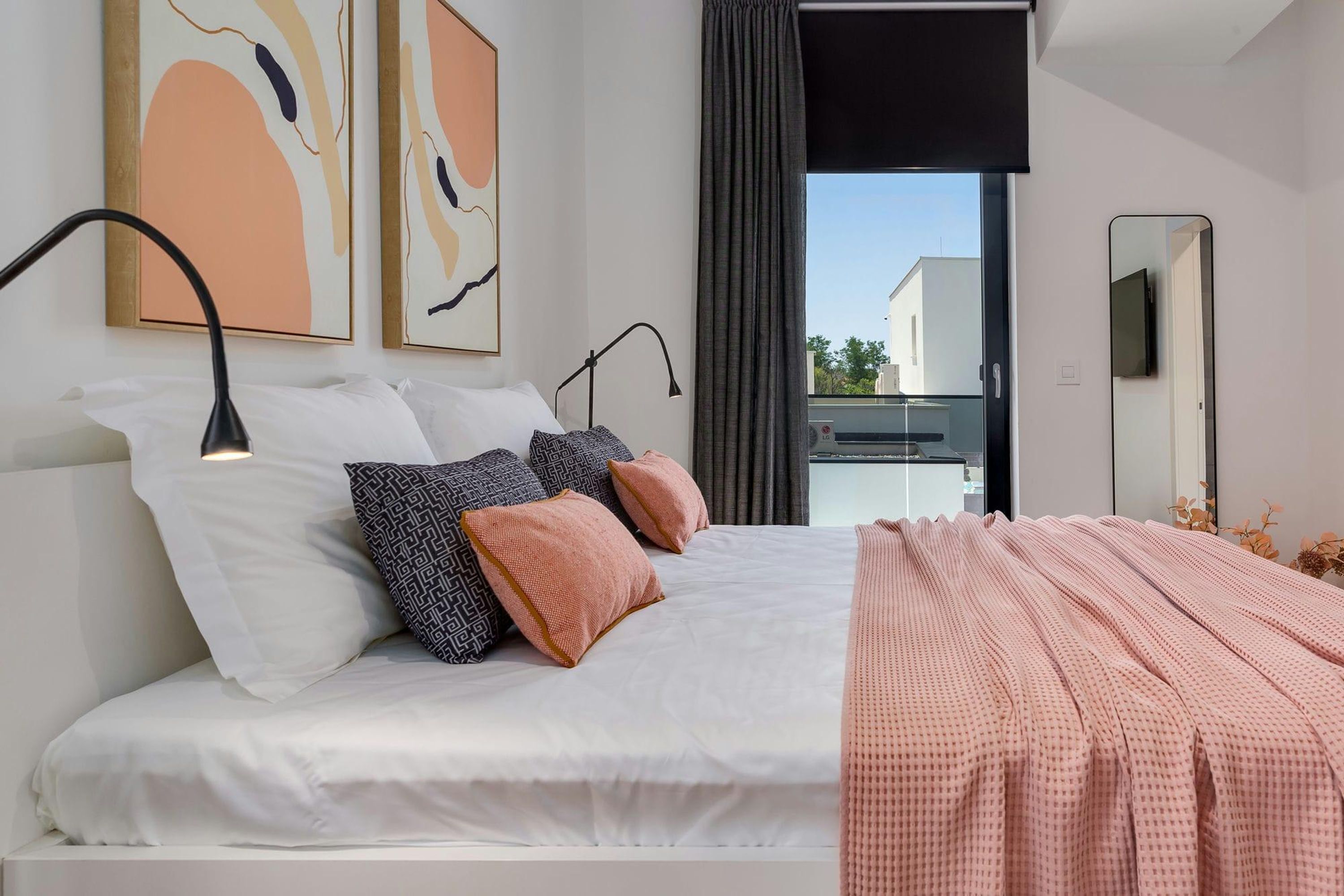 A modern bedroom with a large bed, two bedside lamps, abstract wall art, a balcony with a view, and a mirror on the wall.