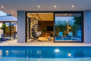 Una casa moderna con piscina, una puerta de cristal que da a un patio con mobiliario exterior, y una silla colgante.