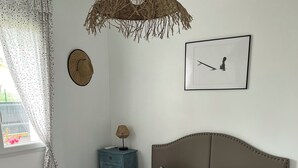 Un lit bien fait, avec une tête de lit beige, un luminaire suspendu en fibres tressées, un tableau encadré accroché au mur et une petite table de chevet avec une lampe.